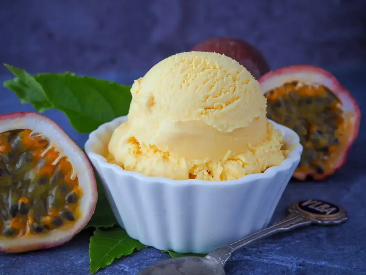 Cremiges Maracuja Eis in einer weißen Schale, umgeben von frischen Passionsfrüchten und grünen Blättern. Ein Löffel liegt daneben.