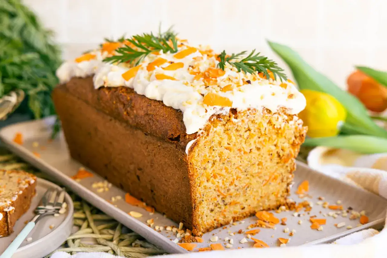 Saftiger karottenkuchen in Kastenform, dekoriert mit Frischkäse-Frosting, gehackten Nüssen und Karottenstückchen.