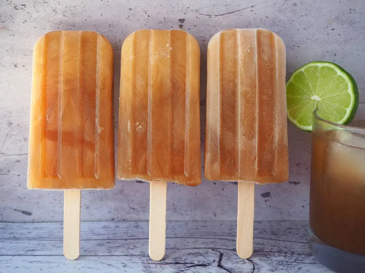 Erfrischendes Cola-Eis selber machen: Drei Eis am Stiel neben einem Glas Cola und einer Limettenscheibe.