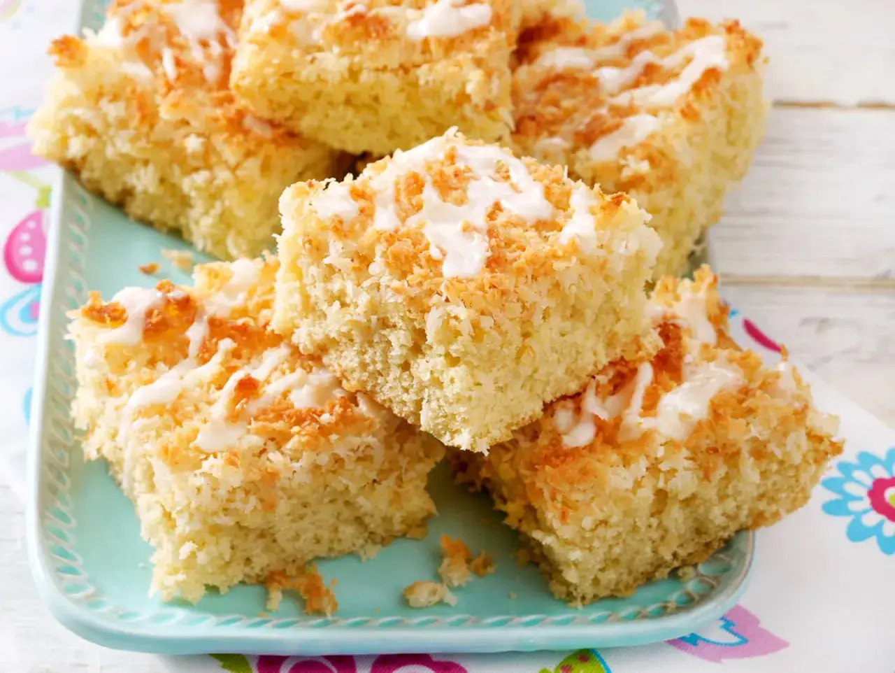 Saftige Stücke Kokos Kuchen mit weißer Glasur und gerösteten Kokosflocken, auf einem türkisfarbenen Teller.