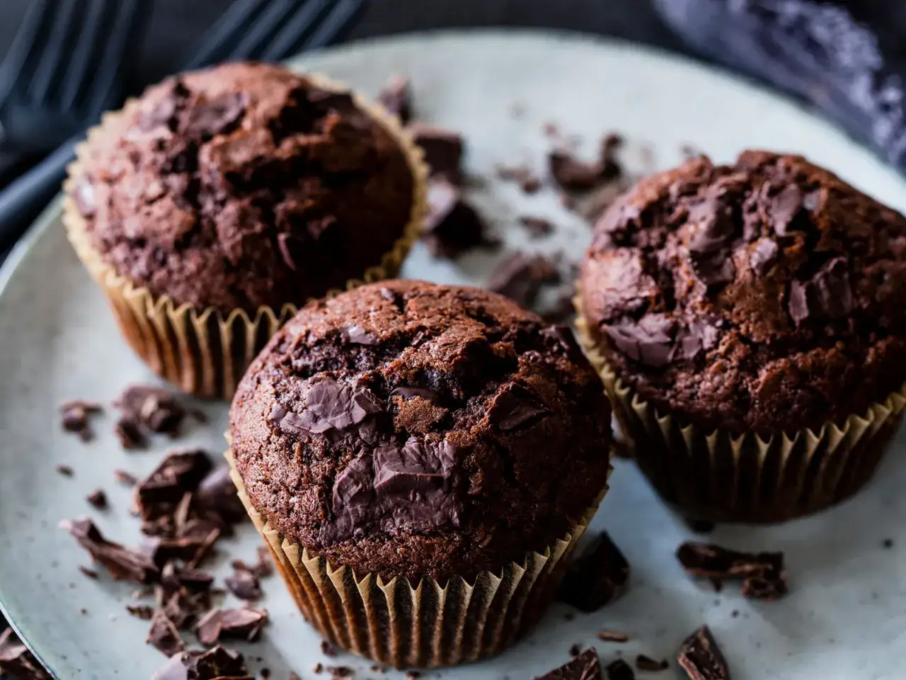 Drei saftige Schoko-Muffins mit Schokoladenstückchen auf einem Teller, umgeben von Schokoladenraspeln. Ein perfektes Muffins Rezept für Genießer.