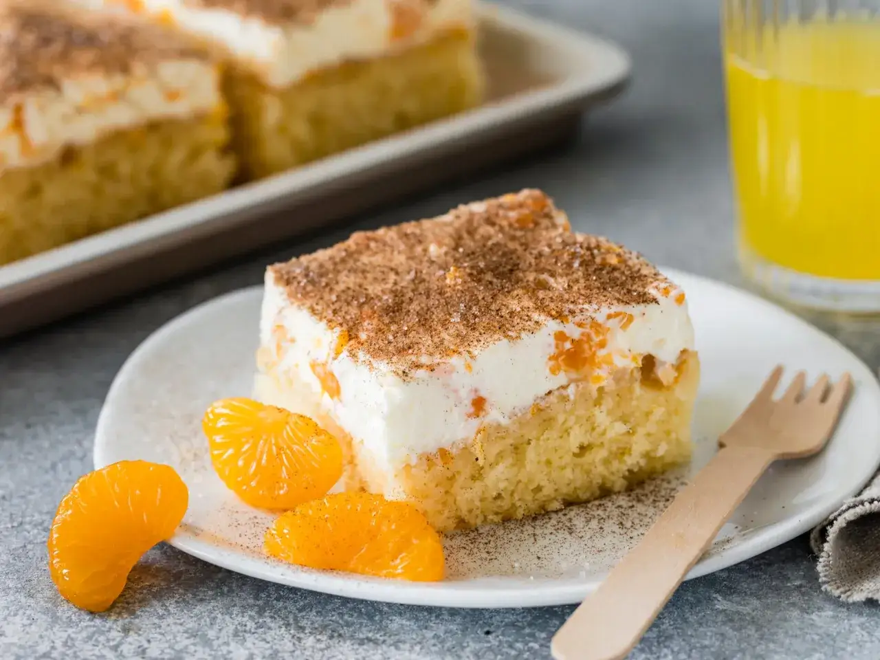 Ein Stück saftiger Fanta Kuchen mit cremigem Topping, Mandarinenstücken und Zimt bestreut. Daneben liegen frische Mandarinenfilets.