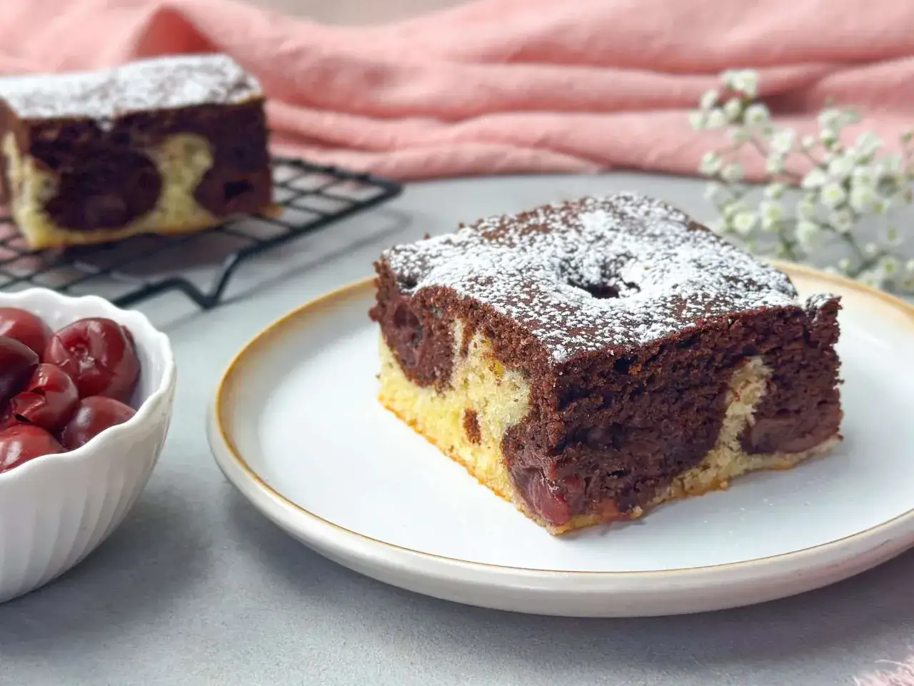 Ein Stück saftiger Marmorkuchen mit Kirschen, bestäubt mit Puderzucker. Daneben eine Schale mit frischen Kirschen.