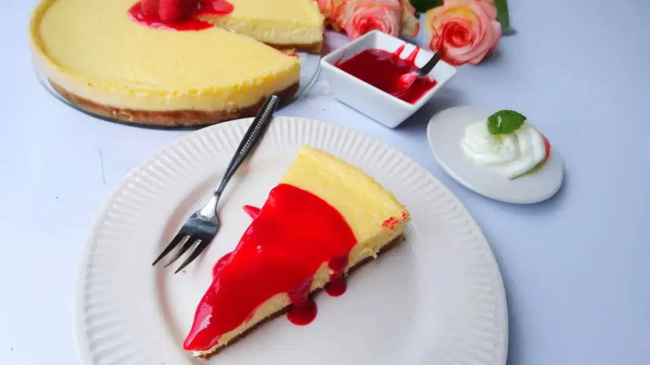 Ein Stück New York Cheesecake mit roter Soße und Himbeeren, daneben eine kleine Schale mit Soße und ein Klecks Sahne mit Minze.