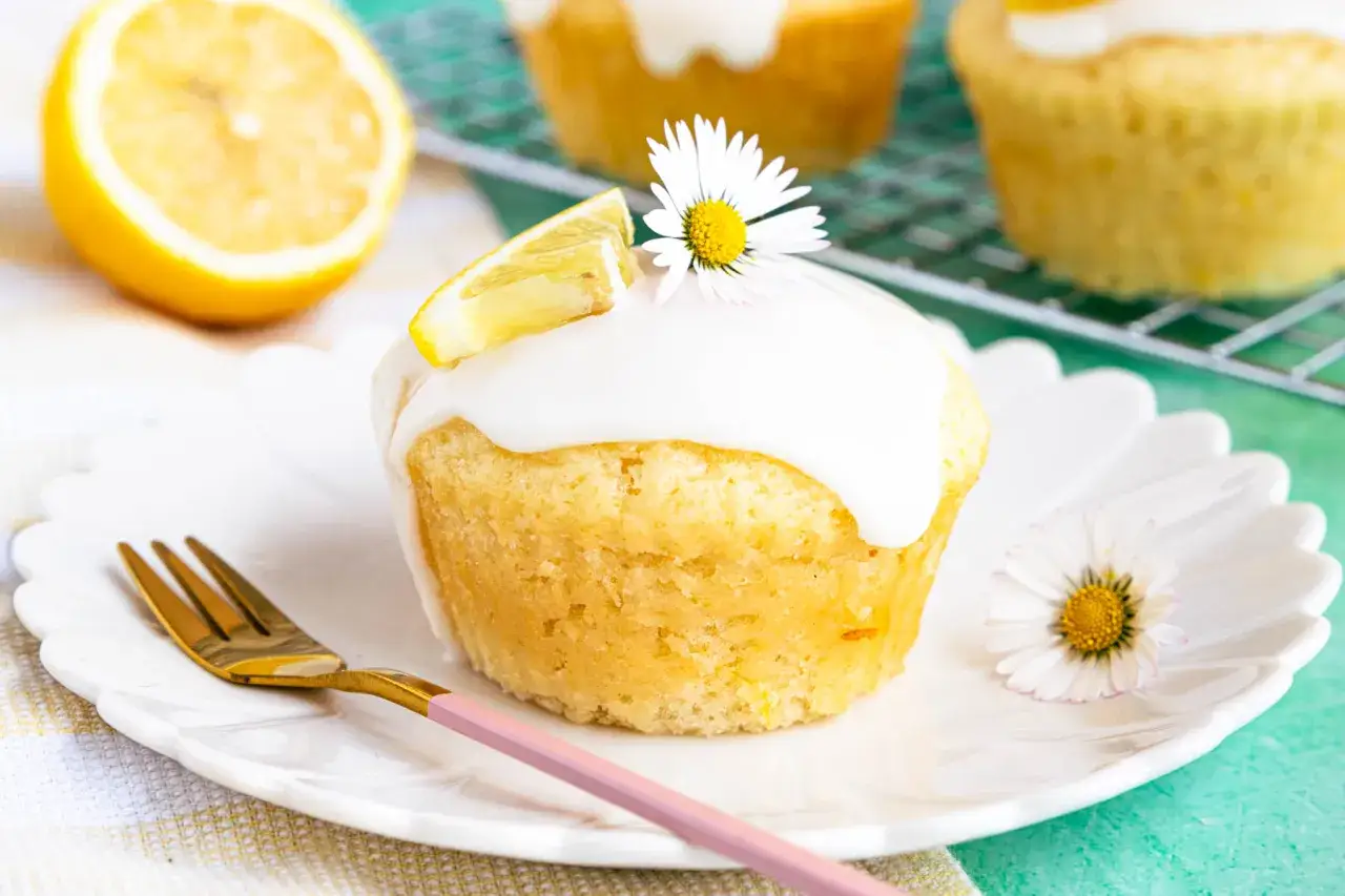Vegane Zitronenmuffins mit Zuckerguss, Zitronenspalte und Gänseblümchen auf einem Teller.