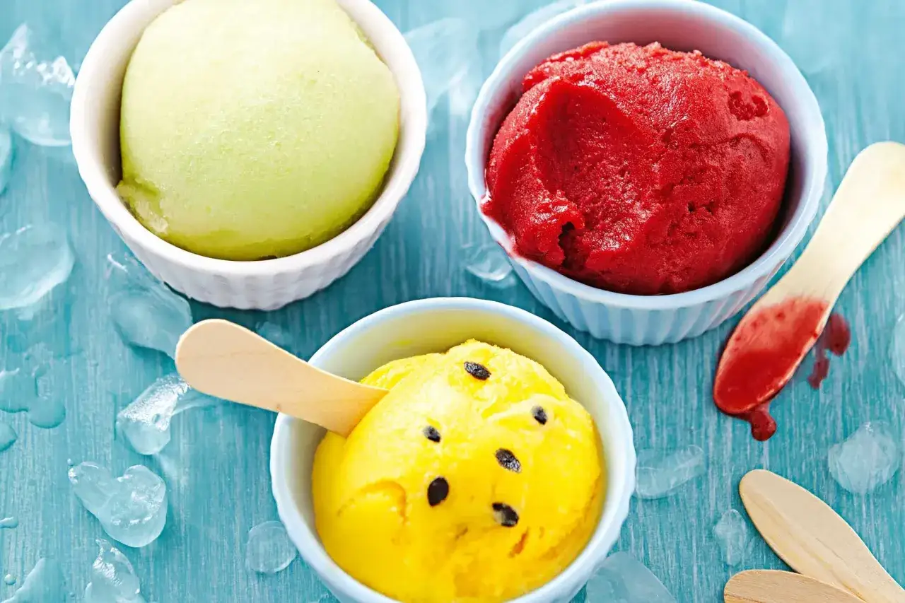 Erfrischende Sorbet-Rezepte: Grünes Melonen-, rotes Beeren- und gelbes Maracuja-Sorbet auf blauem Untergrund mit Eiswürfeln.