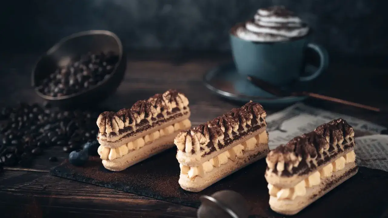 Drei köstliche Tiramisu-Riegel mit Kakaopulver bestäubt, daneben Kaffeebohnen und eine Tasse Cappuccino. Ein perfektes Tiramisu Rezept.