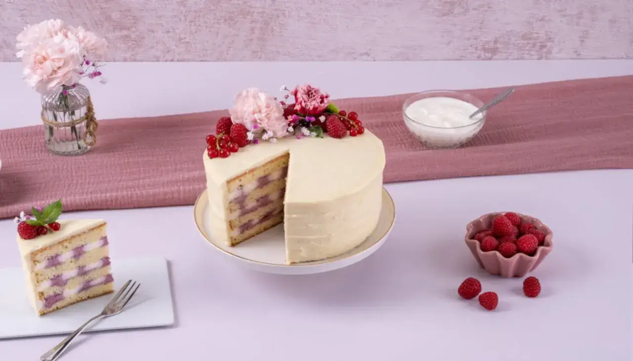 Eine köstliche Himbeer Sahne Torte, dekoriert mit frischen Beeren und Blumen. Ein Stück ist bereits angeschnitten.