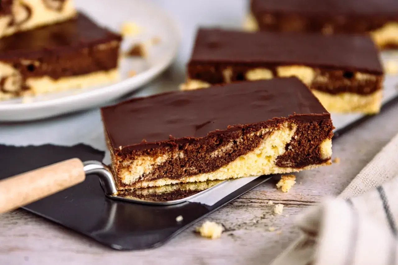 Ein St&uuml;ck saftiger Marmorkuchen vom Blech, mit Schokoladenglasur, wird auf einem Kuchenheber pr&auml;sentiert.