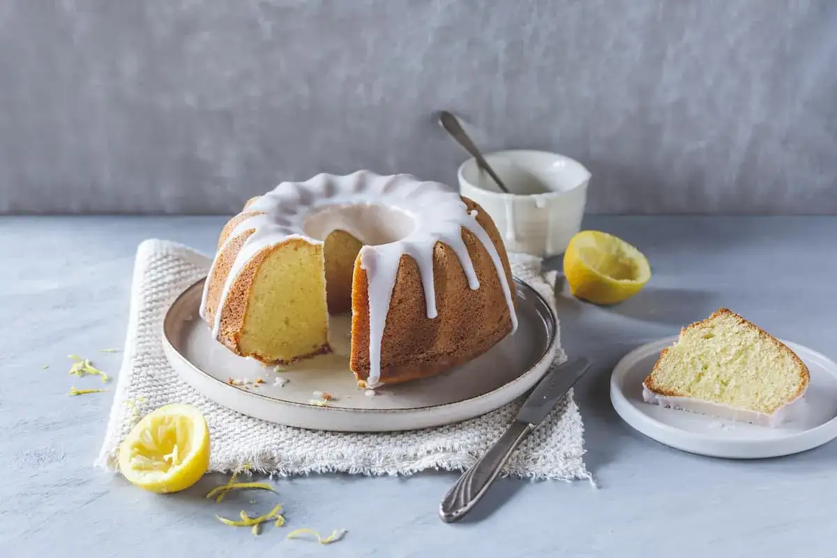 Ein saftiger Zitronen Gugelhupf mit Zuckerguss, daneben eine Zitronenh&auml;lfte und ein St&uuml;ck Kuchen.