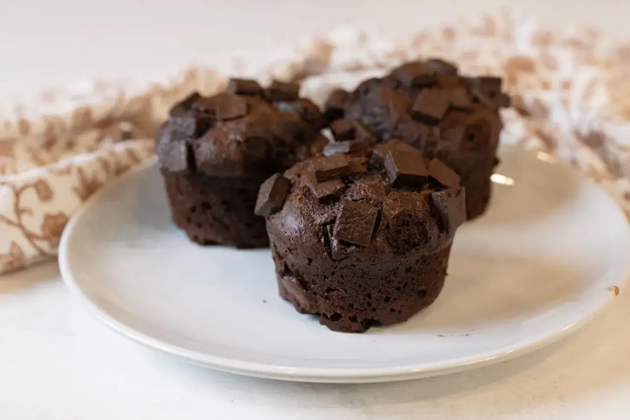Drei saftige, vegane Schokomuffins mit Schokostückchen auf einem weißen Teller, daneben ein gemustertes Tuch.