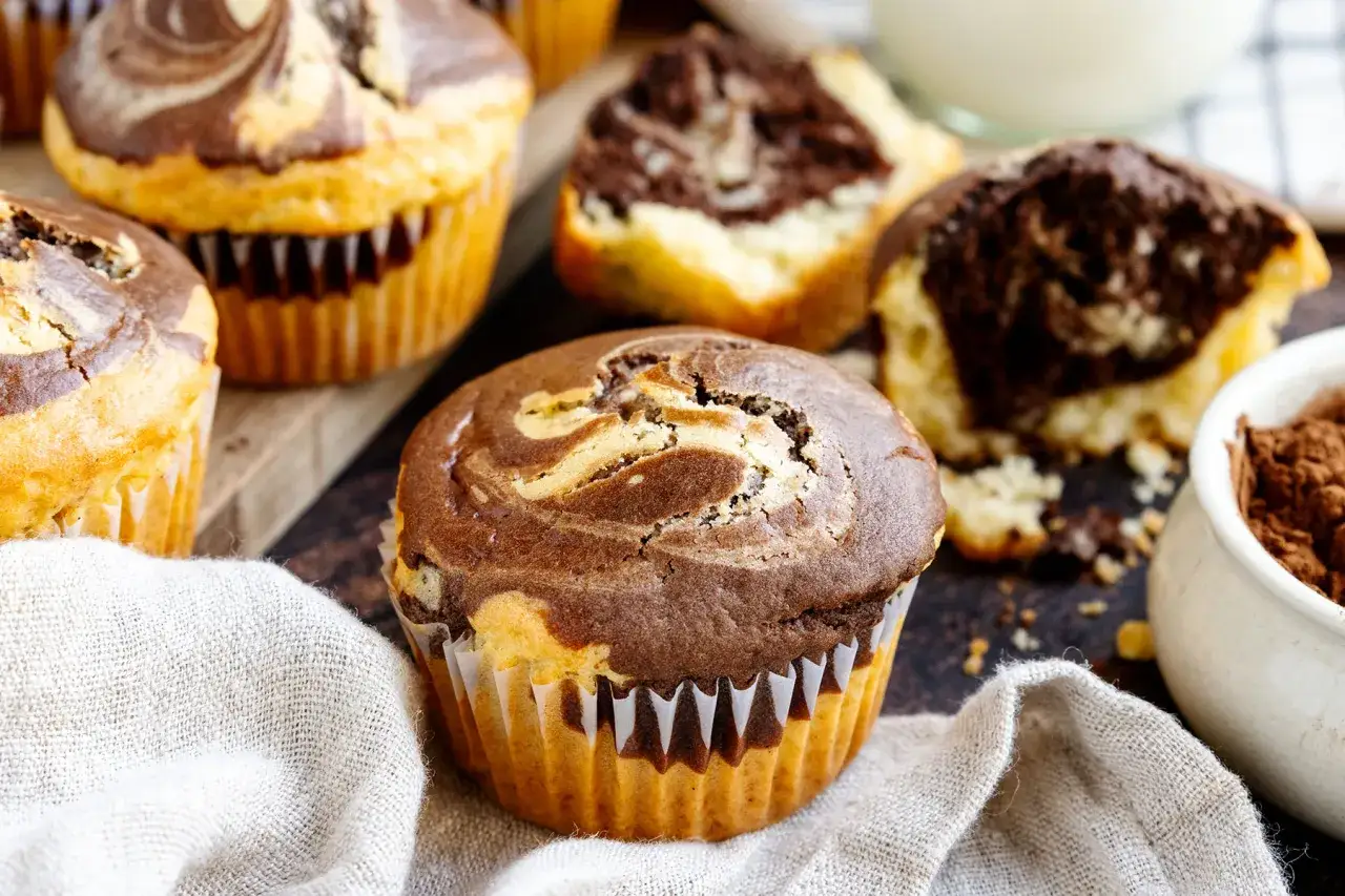 Saftige Marmor Muffins mit Swirls aus hellem und dunklem Teig, auf einem rustikalen Holzbrett, mit einem Stück Stoff und einer Schale Kakaopulver.
