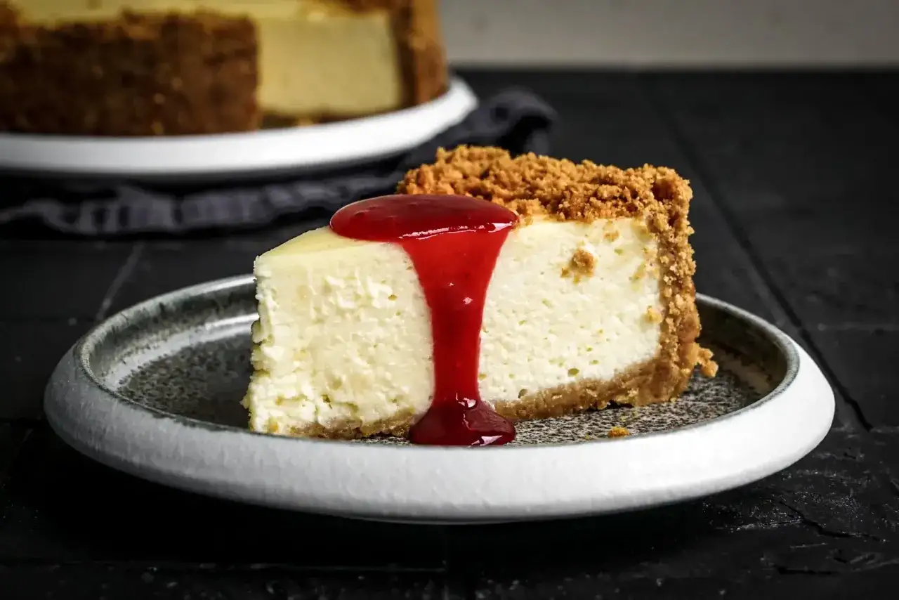 Ein Stück cremiger New York Cheesecake mit Keksboden, übergossen mit roter Fruchtsauce, auf einem grauen Teller.