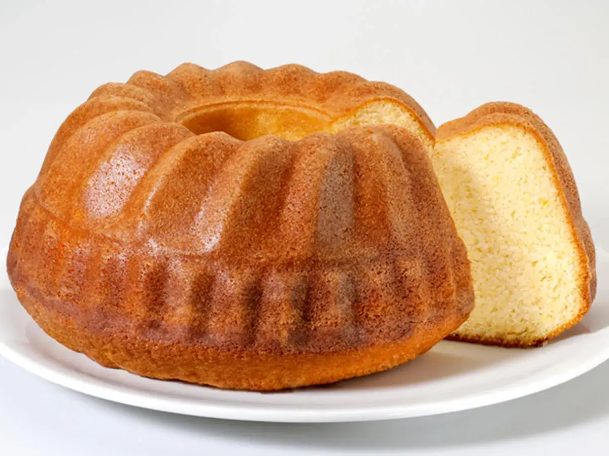 Goldbrauner Gugelhupf, ein Kuchen mit vielen Eiern, auf weißer Platte. Ein Stück ist abgeschnitten und zeigt die lockere Krume.