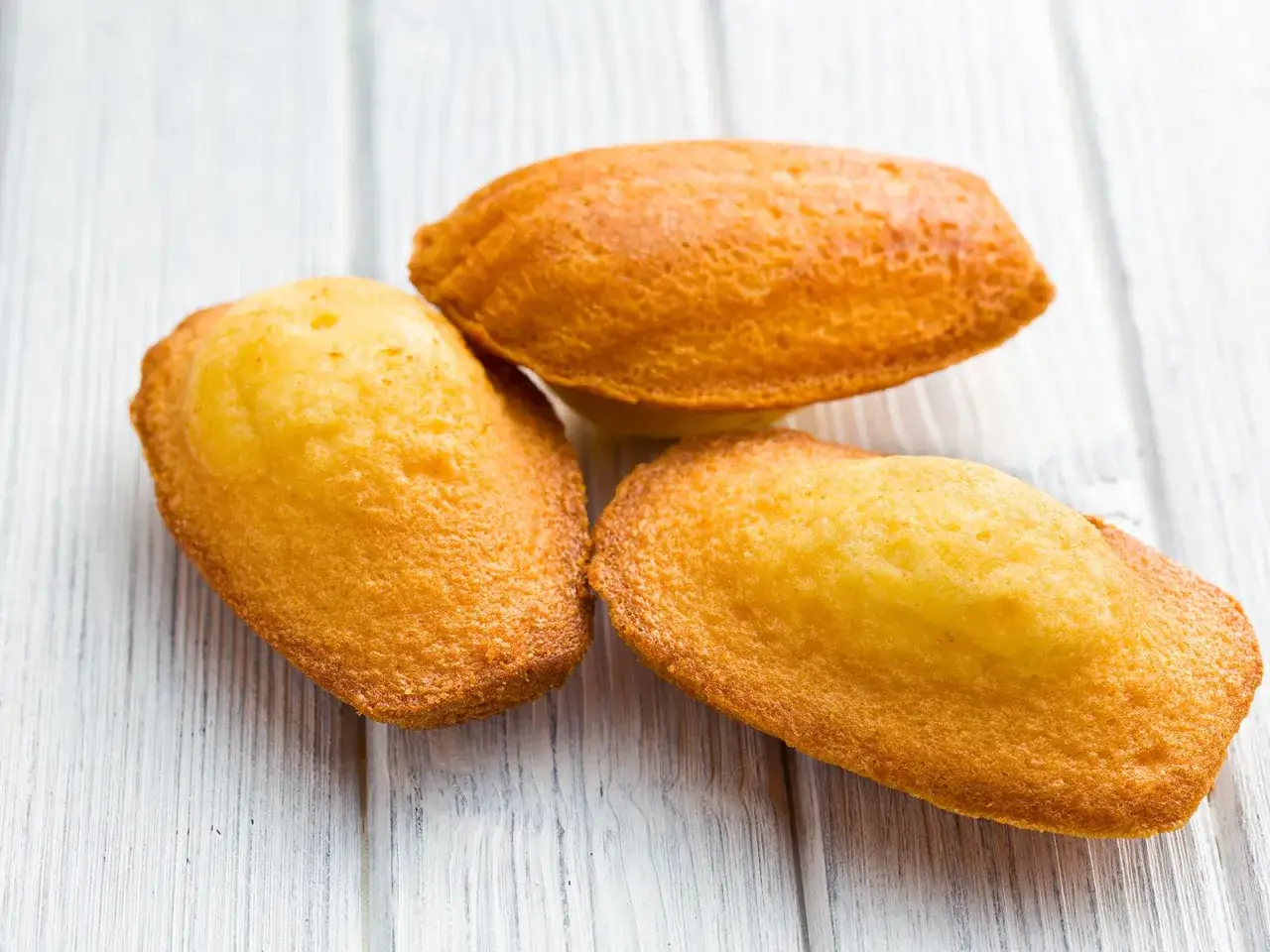 Drei goldbraune Madeleines liegen auf einer weißen Holzoberfläche. Ein köstliches Madeleines Rezept zum Nachbacken.