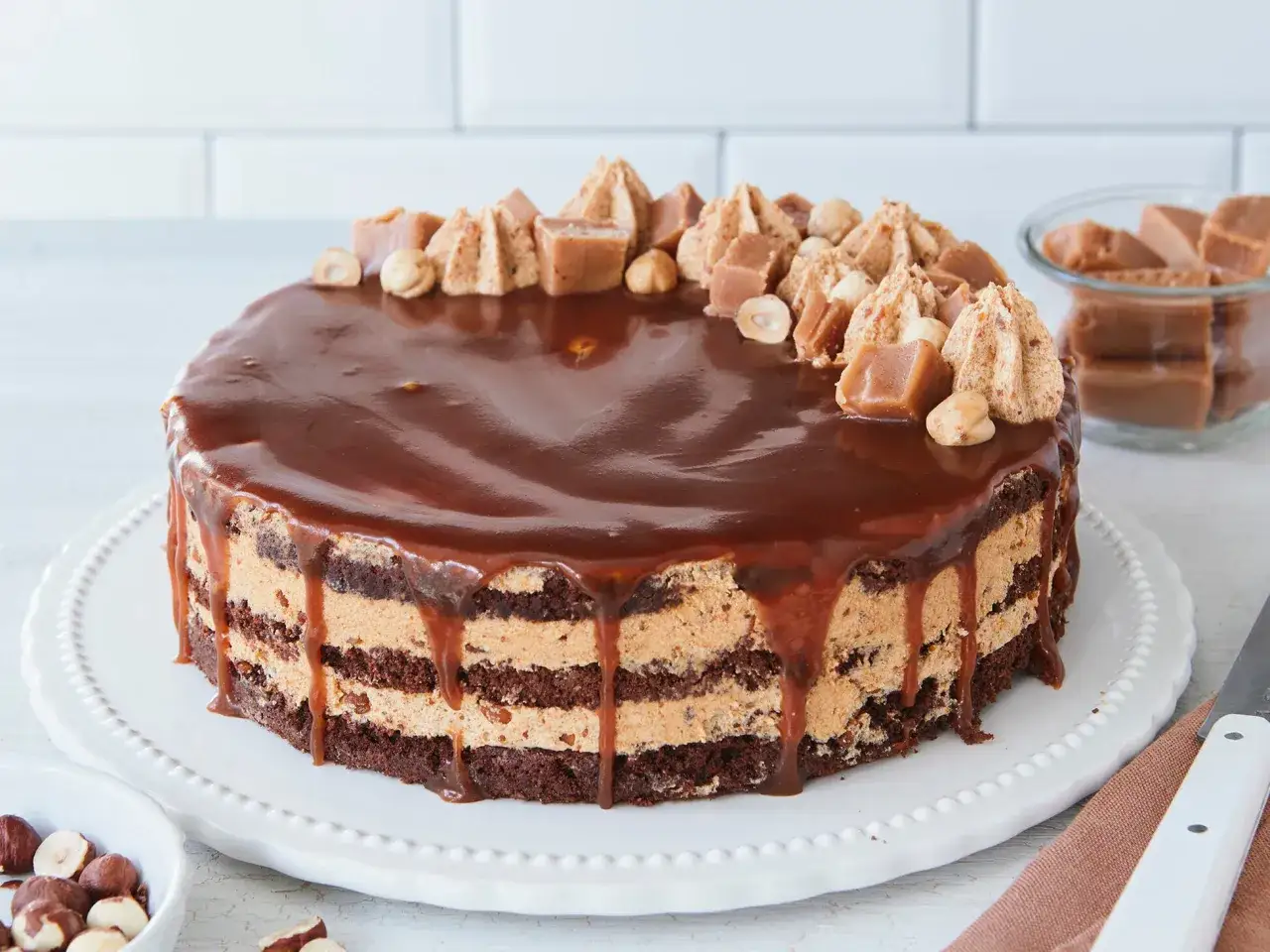 Eine köstliche Maxi King Torte mit Schokoladenböden, Karamellcreme und glänzender Schokoladenglasur, dekoriert mit Karamellstücken und Haselnüssen.