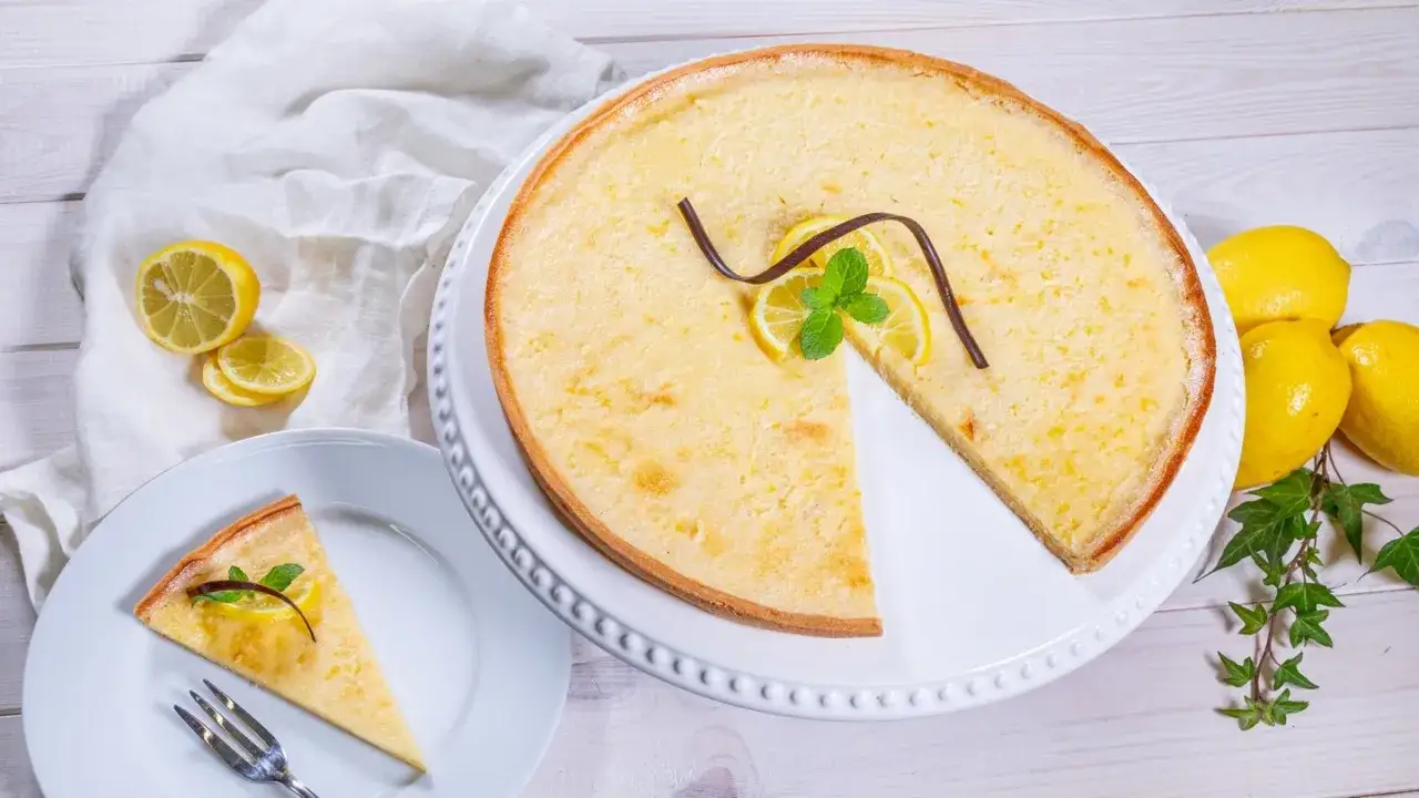 Ein köstliches Zitronentarte Rezept: Ganze Tarte und ein Stück auf Teller, garniert mit Zitronenscheiben, Minze und Schokolade. Frische Zitronen liegen daneben.