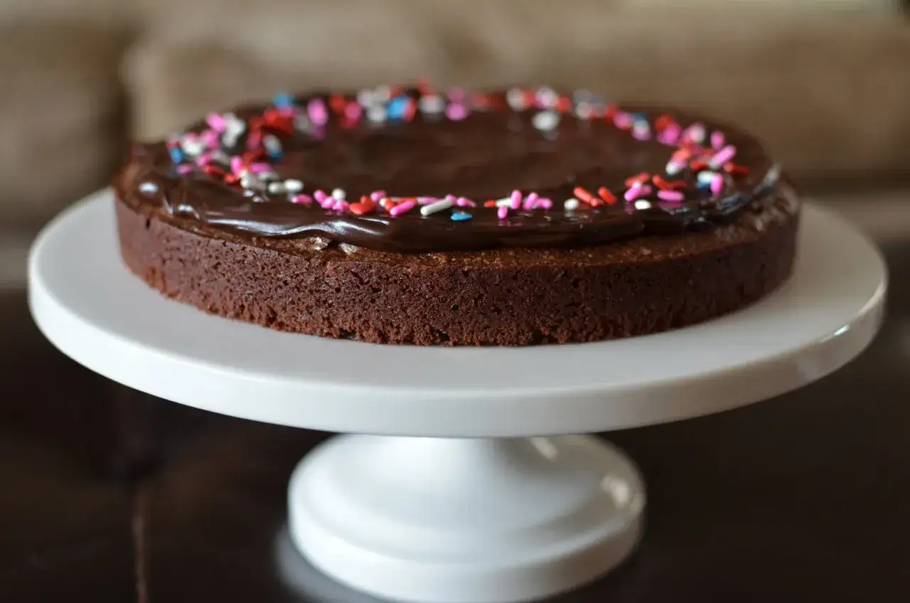 Schokoladenkuchen mit Schokoladenglasur und bunten Streuseln auf einem weißen Kuchenständer.