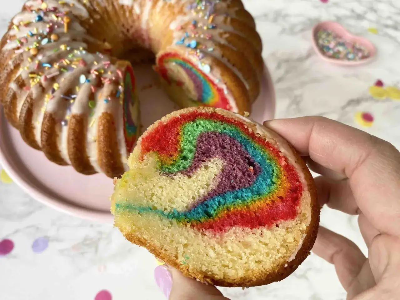 Ein St&uuml;ck eines bunten Regenbogenkuchens mit Zuckerguss und Streuseln wird von einer Hand gehalten. Der Kuchen hat leuchtende Farben im Inneren.