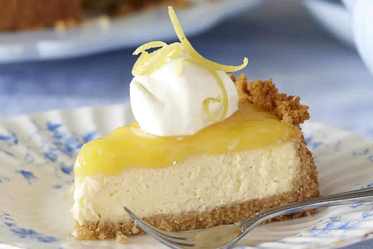Ein Stück cremiger Lemon Cheesecake mit Sahnetupfer und Zitronenzesten auf einem blauen Blumenmuster-Teller.