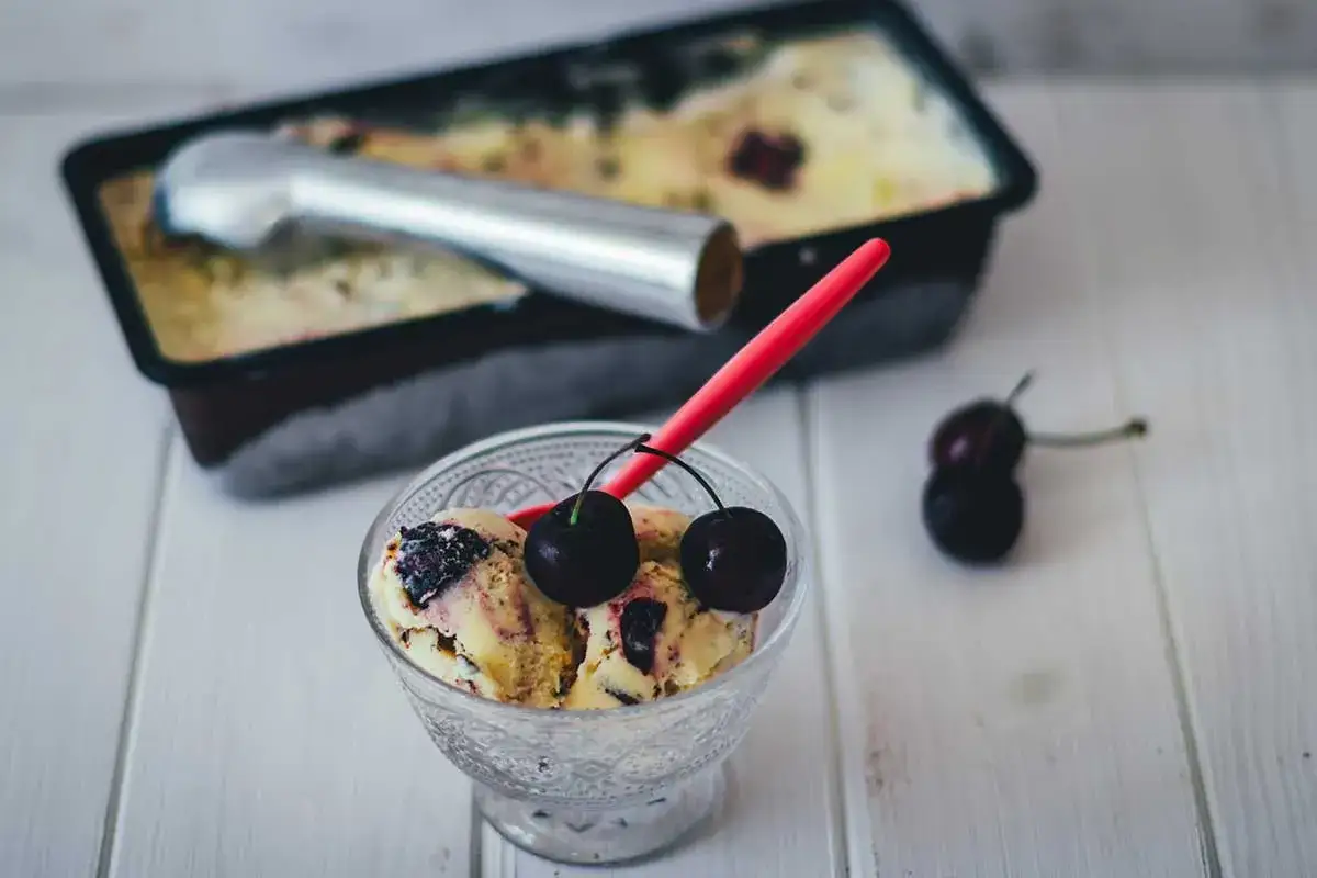 Schüssel mit Kirsch Eis, garniert mit frischen Kirschen. Im Hintergrund eine Eisschale mit Eisportionierer.