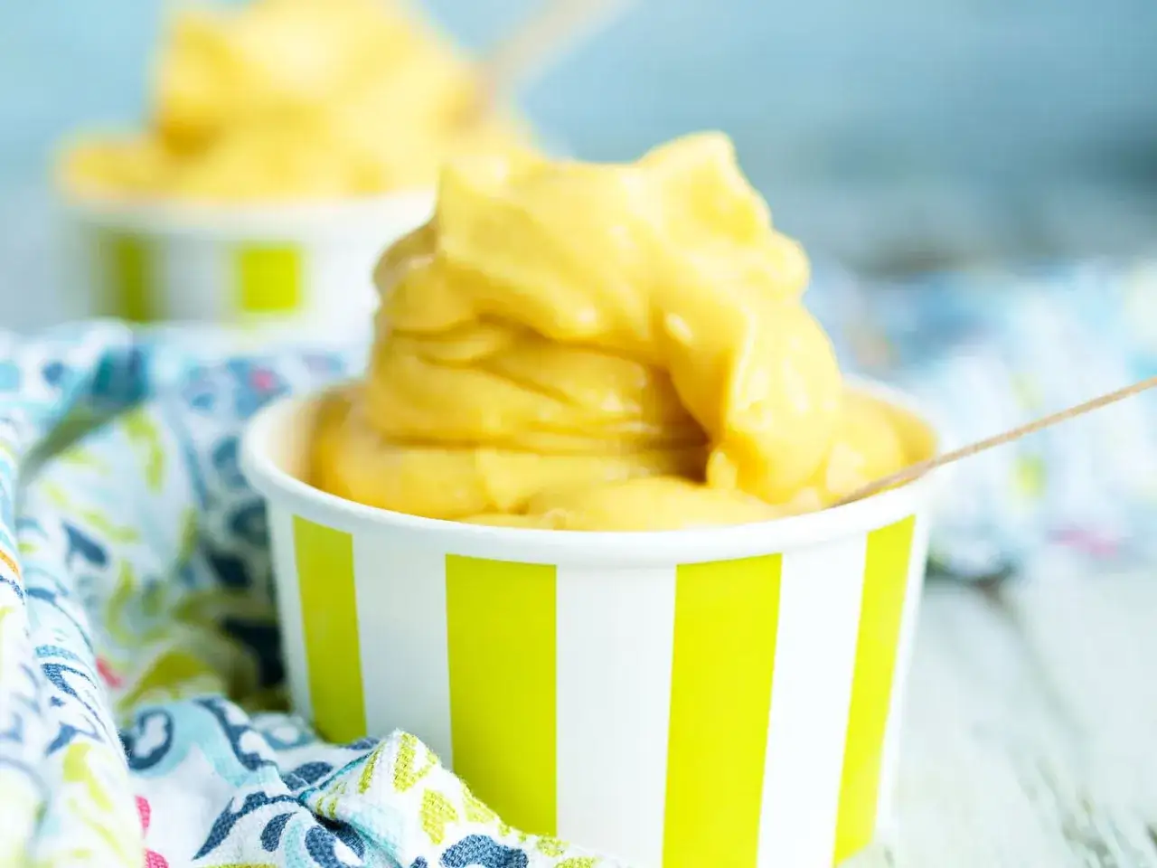 Cremiges Mango Eis in einem gestreiften Becher, daneben ein bunter Stoff. Perfekt für heiße Tage!