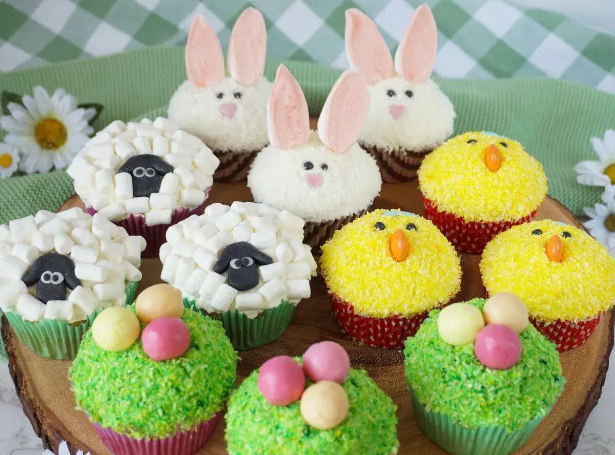 Bunte Oster Cupcakes: süße Lämmchen, Küken und Hasen auf einem Holzteller, dekoriert mit Marshmallows, Kokosraspeln und bunten Eiern.
