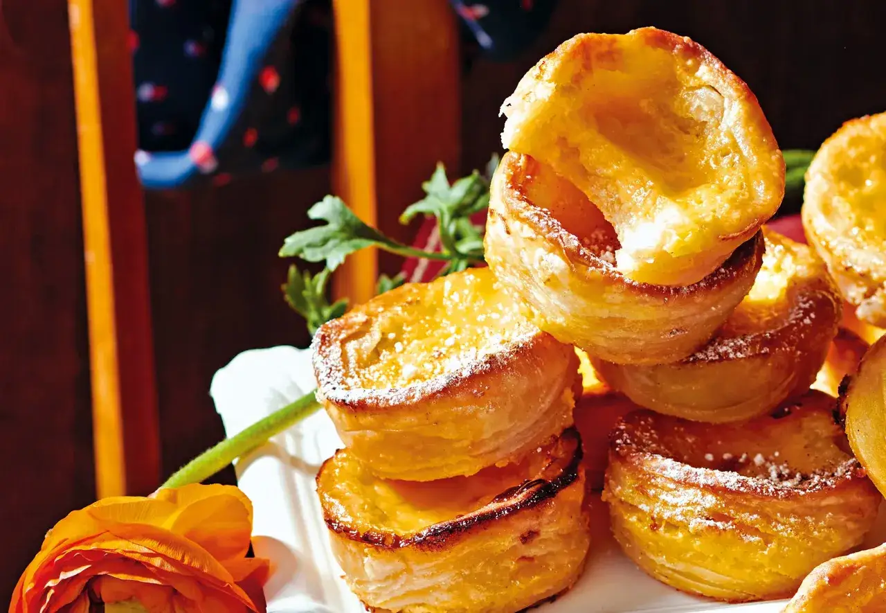 Gestapelte, goldbraune Pastel de Nata mit Puderzucker bestäubt, daneben eine orangefarbene Ranunkel.