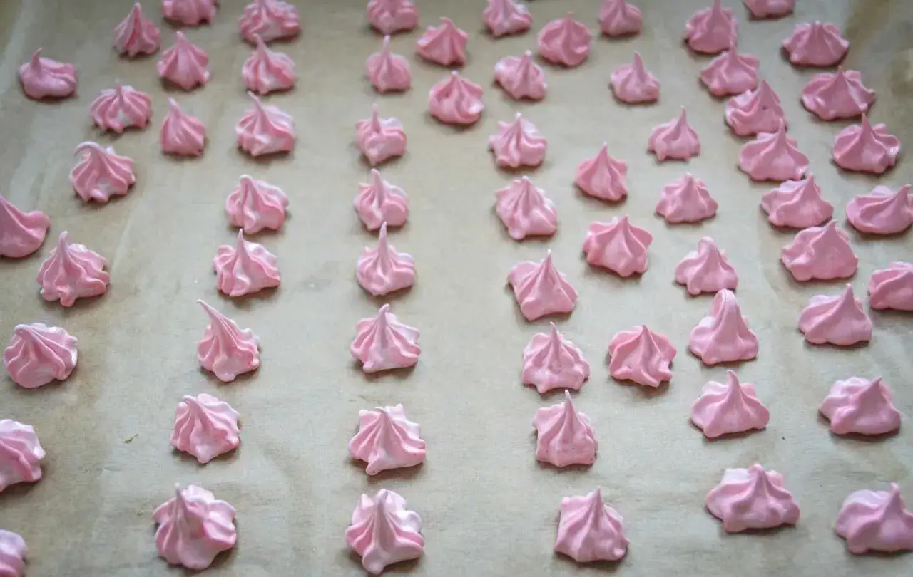 Viele kleine, rosafarbene Meringues liegen auf Backpapier. Ein einfaches Meringues Rezept für süße Leckereien.