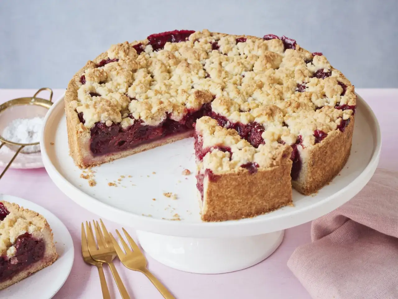 Ein saftiger Kirschkuchen mit knusprigen Streuseln auf einem wei&szlig;en Tortenst&auml;nder. Ein St&uuml;ck ist bereits herausgeschnitten.