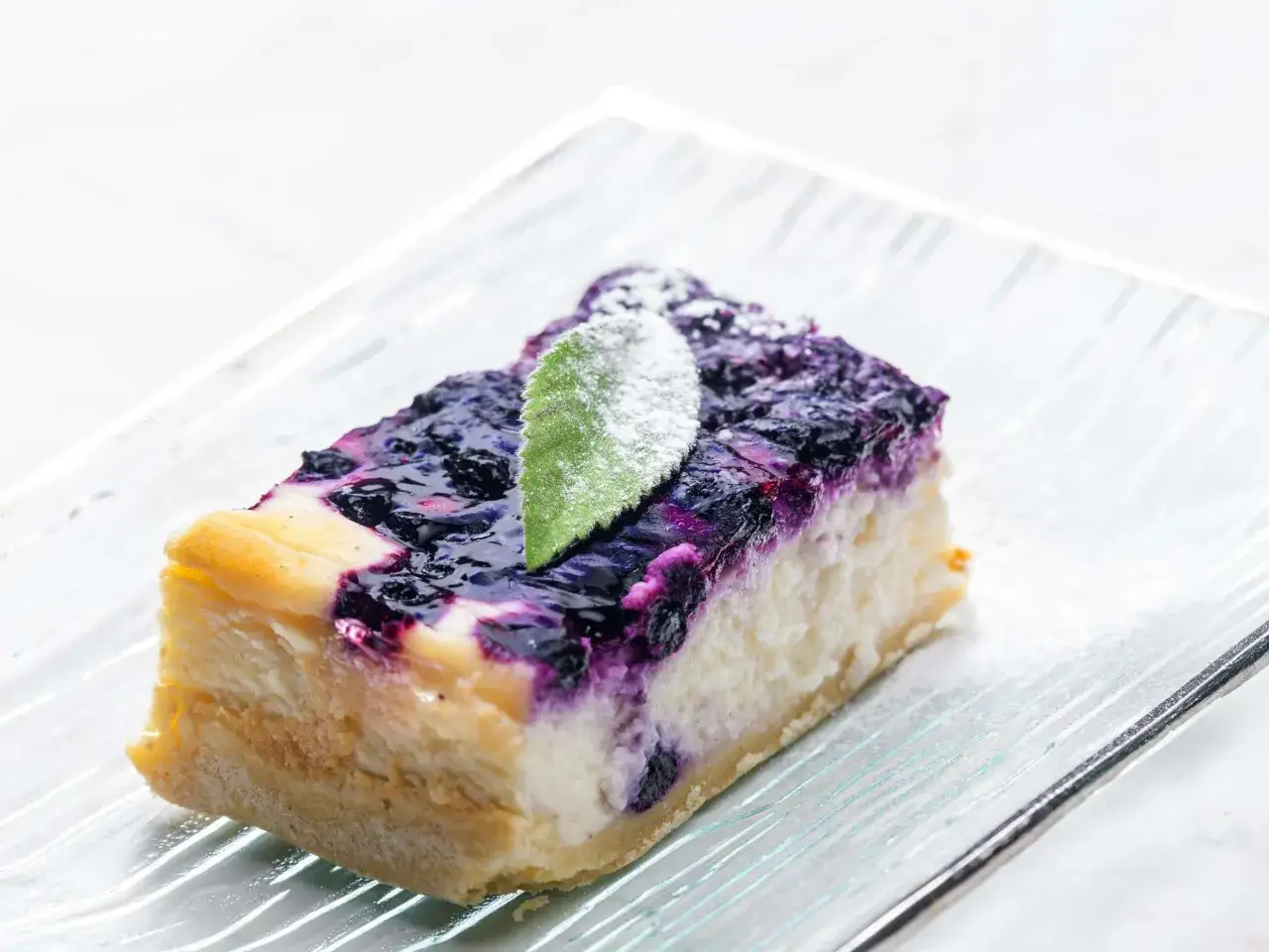 Ein Stück cremiger Blueberry Cheesecake mit Blaubeersauce und einem grünen Blatt, bestäubt mit Puderzucker.