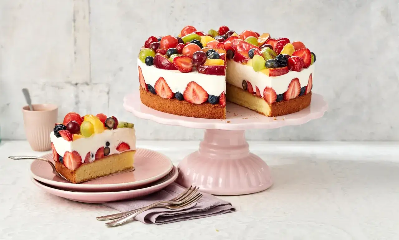 Fruchtige Torte ohne Backen, dekoriert mit Erdbeeren, Blaubeeren, Trauben und Pfirsichen auf einer cremigen Füllung. Ein Stück ist bereits auf einem Teller angerichtet.