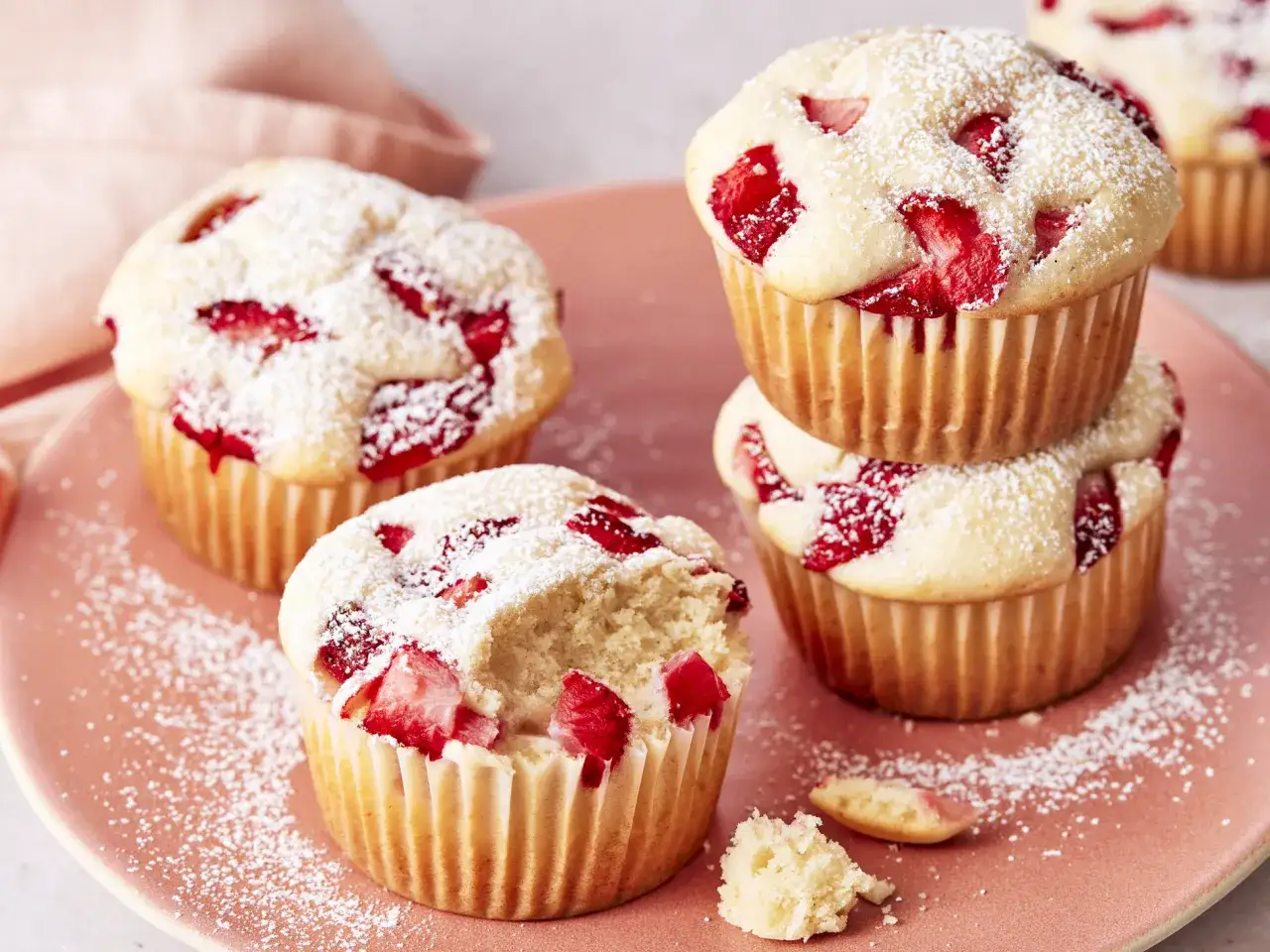 Saftige Erdbeer Muffins, mit Puderzucker bestäubt, auf einem rosafarbenen Teller. Ein Muffin ist angeknabbert.