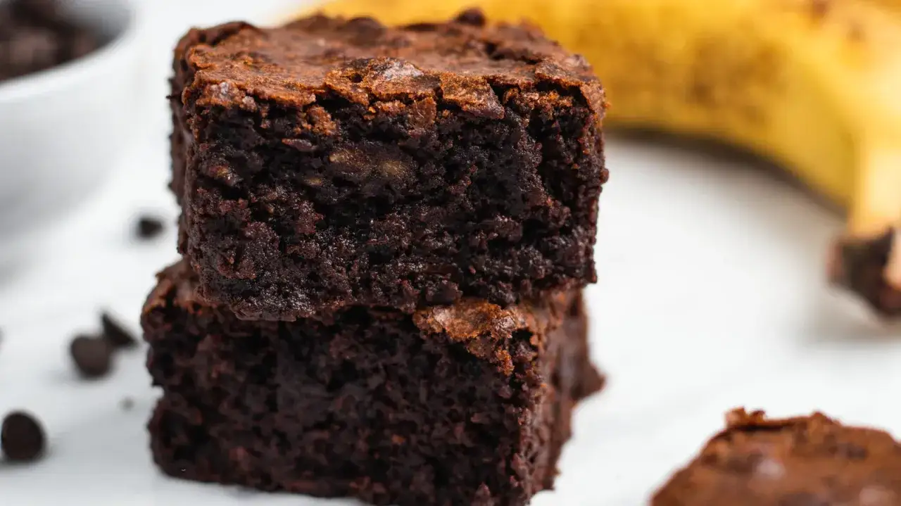 Gestapelte, saftige Bananen-Brownies mit Schokostückchen und einer Banane im Hintergrund.