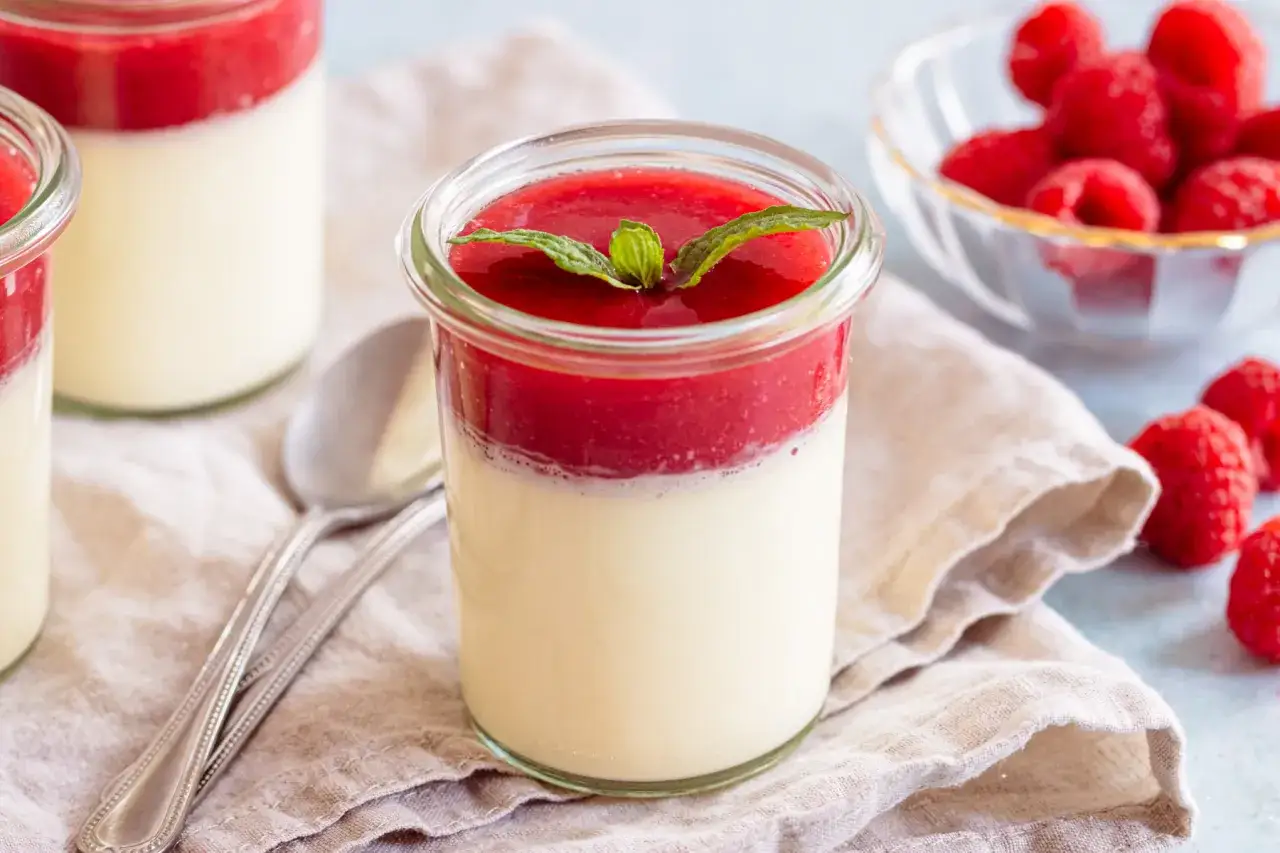 Cremiges Panna cotta im Glas, gekrönt mit fruchtigem Himbeermark und frischer Minze. Daneben ein Löffel und frische Himbeeren.