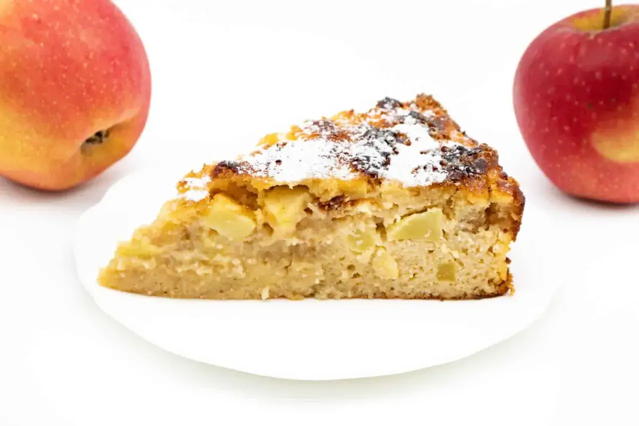 Ein Stück saftiger Apfelkuchen, bestäubt mit Puderzucker, daneben zwei Äpfel. Perfekt als diabetiker kuchen.