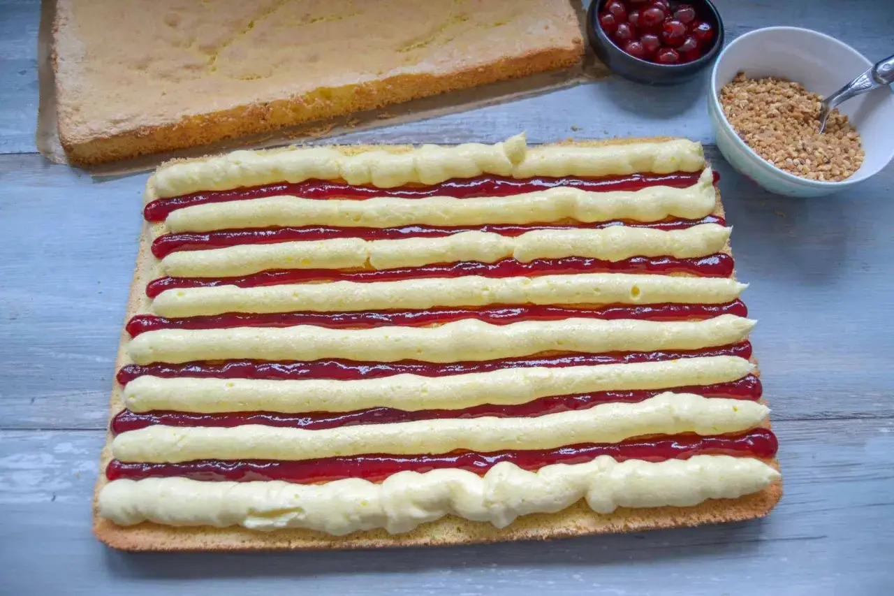 Biskuitboden mit Sahne und roter Marmelade für Frankfurter Kranz Schnitten belegt. Daneben liegen Kirschen und gehackte Nüsse.