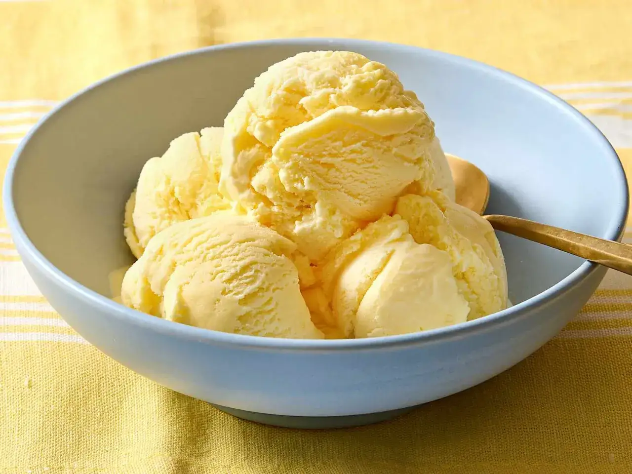 Ein hellblaues Schälchen mit cremigem Zitronen-Eis, das auf einem gelben, gestreiften Tuch steht. Ein goldener Löffel liegt daneben.