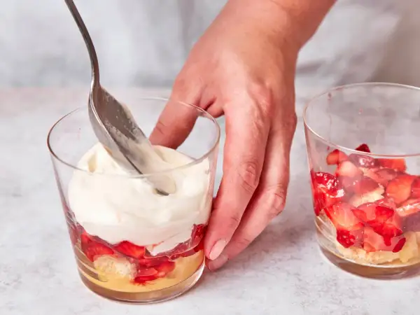 Eine Hand schichtet mit einem Löffel cremiges Topping auf ein Erdbeer-Tiramisu im Glas.