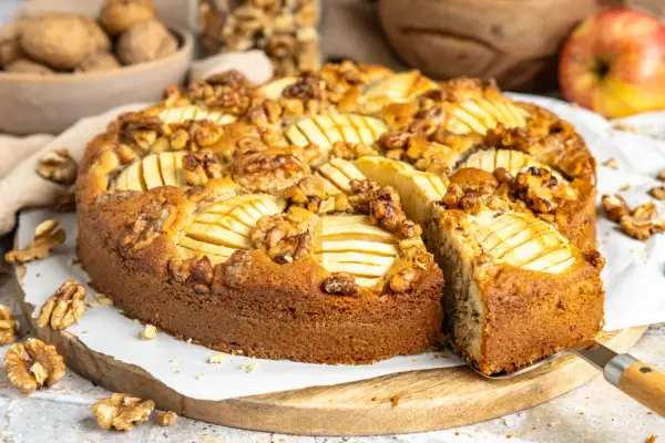 Ein saftiger Apfel Walnuss Kuchen, dekoriert mit Apfelscheiben und ganzen Walnüssen, wird auf einem Holzbrett präsentiert.