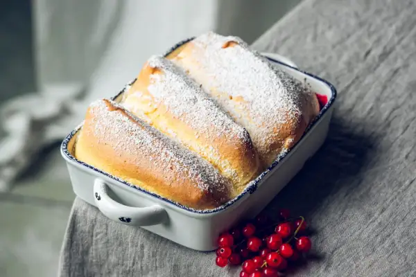 Drei goldbraune Salzburger Nockerl, mit Puderzucker bestäubt, in einer Auflaufform, daneben rote Johannisbeeren.