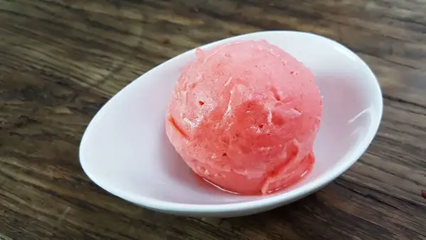Ein Löffel cremiges Erdbeereis mit Sahne in einer weißen Schale auf Holzhintergrund.