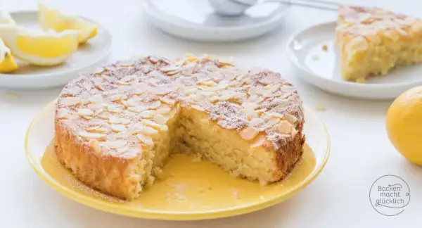 Saftiger mandelkuchen rezept, bestäubt mit Puderzucker und Mandelsplittern, daneben Zitronenspalten und eine ganze Zitrone.