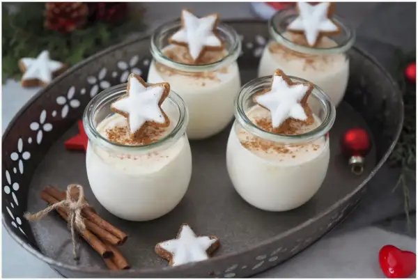 Vier Gläser mit cremigen Parfait-Rezepten im Glas, garniert mit Zimt und Sternenplätzchen. Weihnachtliche Deko mit Zimtstangen und Kugeln.