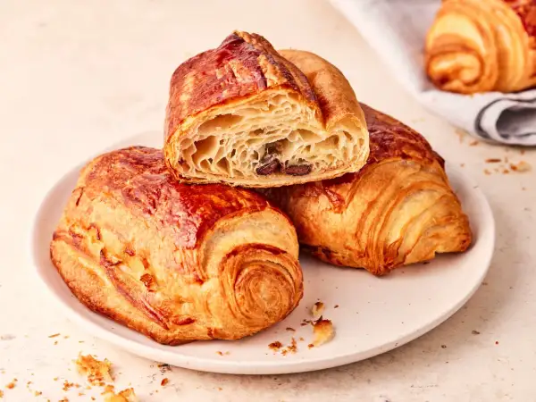 Złociste, chrupiące pain au chocolat na talerzu, jedno przekrojone, ukazujące puszyste wnętrze z kawałkami czekolady.