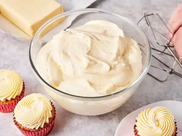 Schüssel mit cremiger amerikanischer Buttercreme, daneben ein Block Butter, Cupcakes und ein Handmixer.