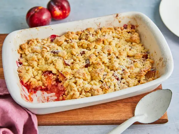 Ein frisch gebackener pflaumen crumble in einer weißen Auflaufform, bereit zum Servieren. Daneben liegen zwei reife Pflaumen.