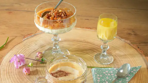 Ein Dessert im Glas mit Rhabarberkompott und Streuseln, daneben ein gelbes Getränk und ein Kaffee.