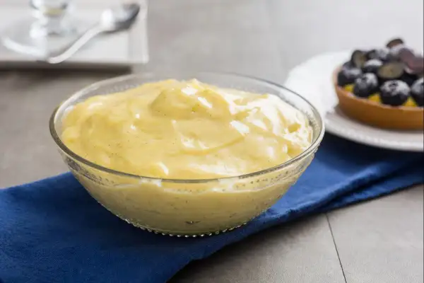 Cremige Crema Pasticcera in einer Glasschüssel, verfeinert mit Vanille. Daneben ein Törtchen mit Blaubeeren.