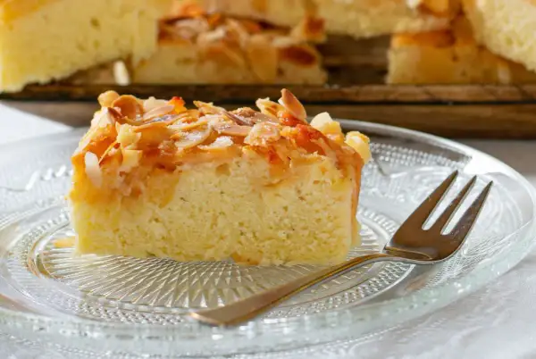 Ein saftiges Stück mandelkuchen vom blech, bestreut mit gerösteten Mandeln und Zucker, liegt auf einem Glas-Teller.